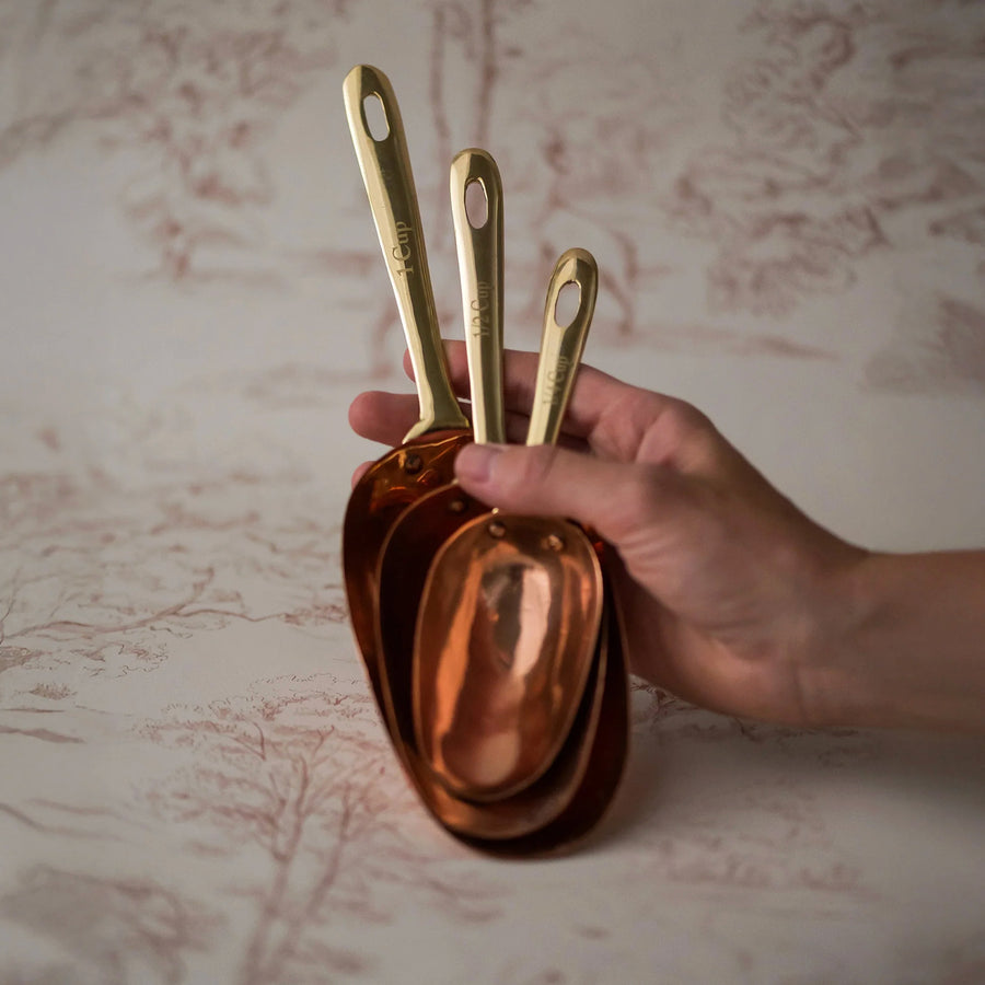 Hand holding three copper measuring spoons against a textured beige background