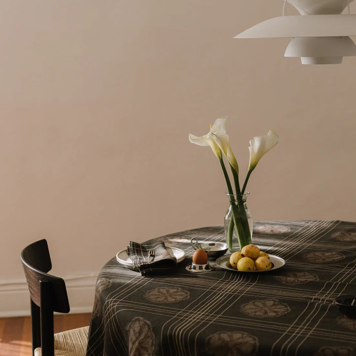 Dining table with a vase of flowers and fruit on a patterned tablecloth against a beige wall.