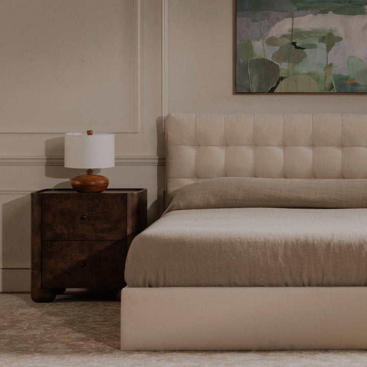 Beige upholstered bed with wooden nightstand and lamp in a bedroom setting.