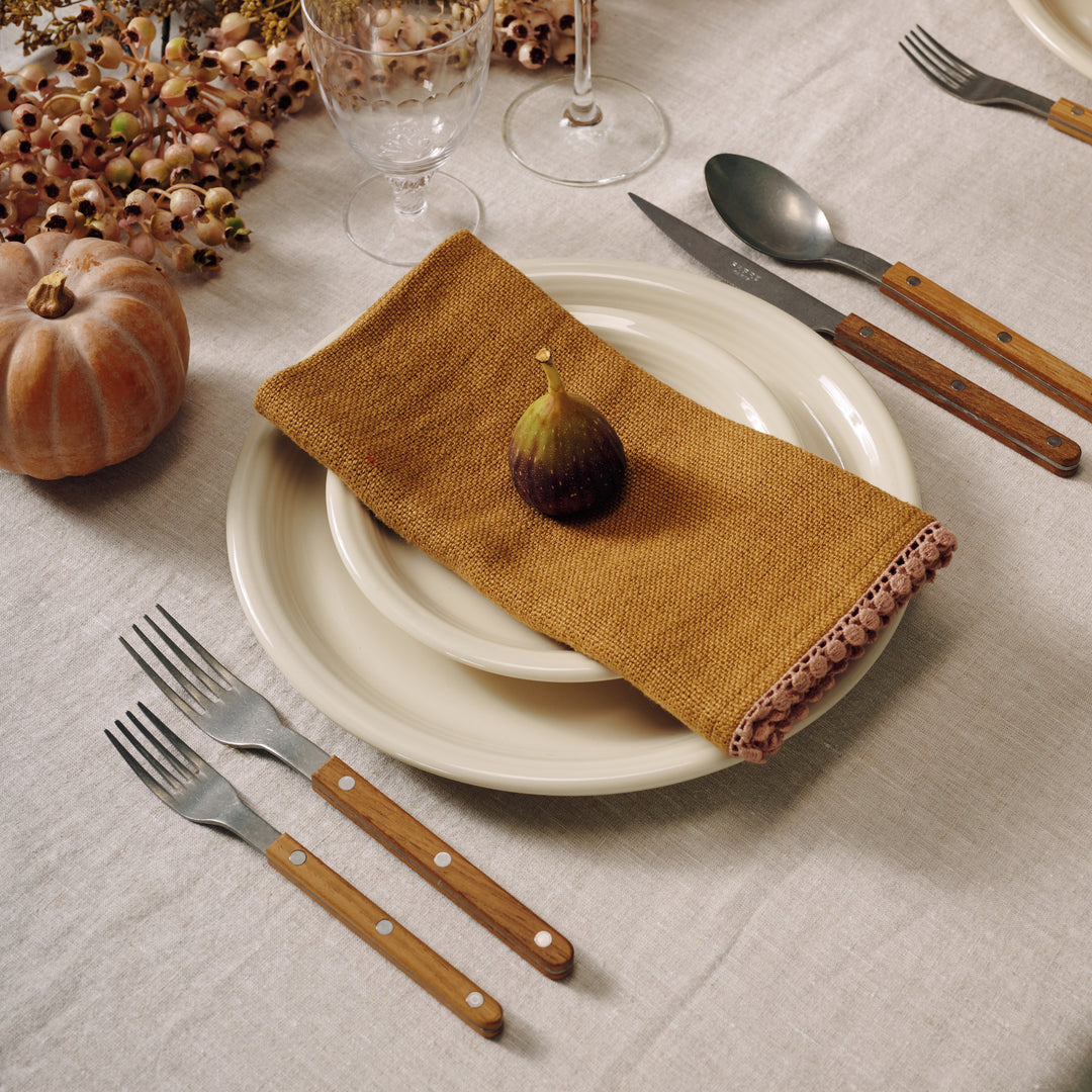 Table setting with white plates, gold napkins, wooden cutlery, and a small fig on a neutral background.