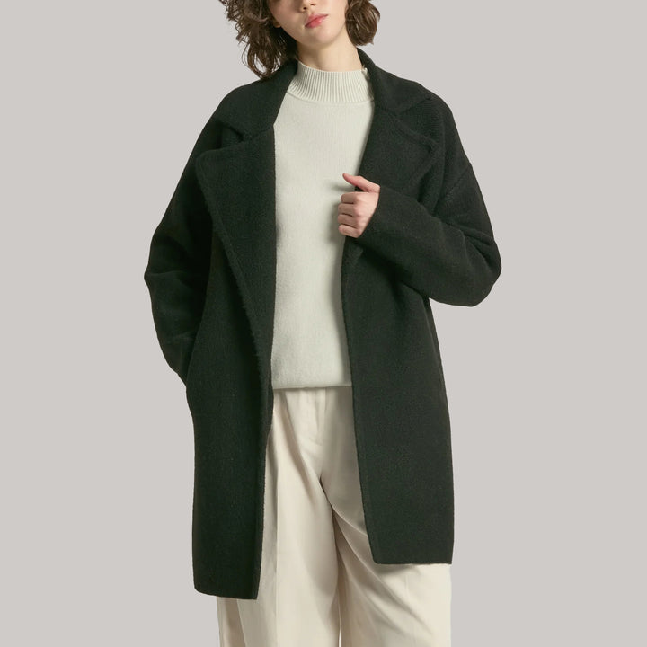 Person wearing a dark black coat over a light sweater and beige pants on a plain background