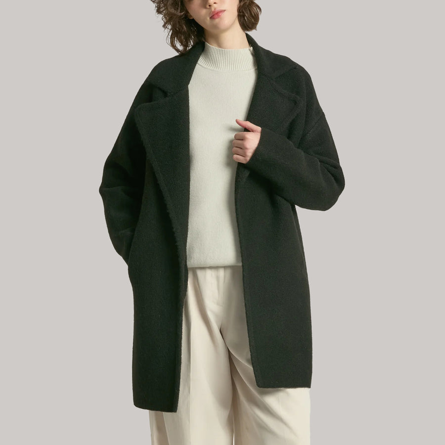 Person wearing a dark black coat over a light sweater and beige pants on a plain background