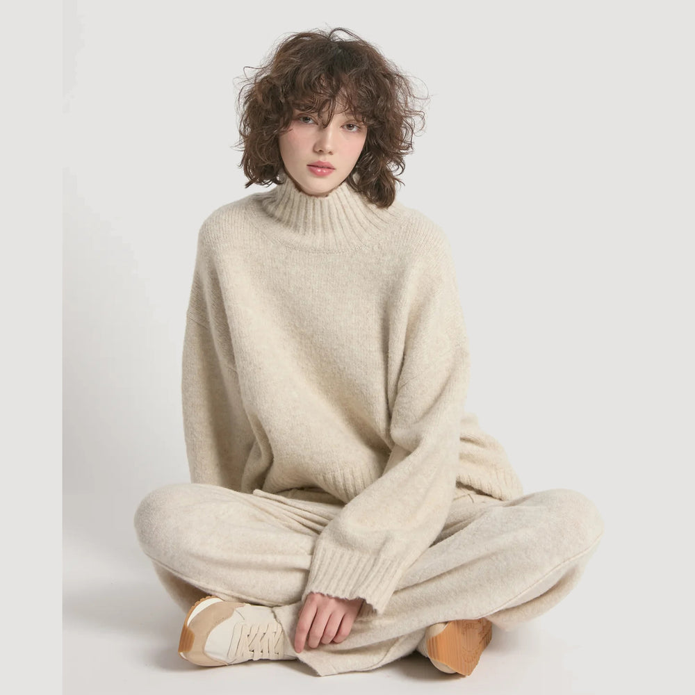 Person wearing a beige sweater and pants sitting on a white background