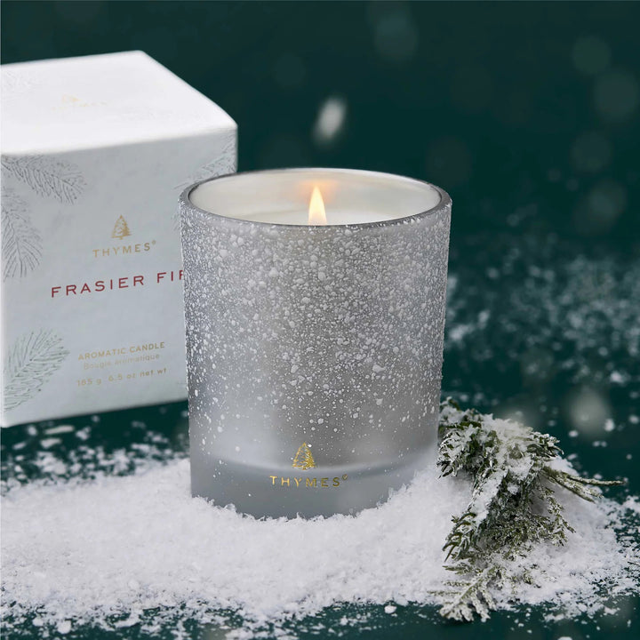 Thymes Frasier Fir candle in a textured glass container with snow and evergreen branches on a dark background